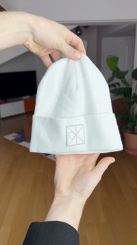 Bonnet blanc côtelé avec un revers plié, présentant un patch logo texturé de forme rectangulaire. Le matériau semble doux et chaud.