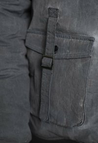 Grijze jas met een getextureerde, matte stof, een groot zakje en een gesp aan de voorkant, met de nadruk op functioneel ontwerp.