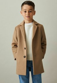 Brun uldblandingsfrakke med skræddersyet design, klippede lapels, to knapper og sidelommer, båret over en beige sweater og blå jeans.