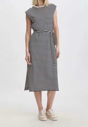 Kobieta ubrana w bezrękawnikową, granatowo-białą sukienkę w paski o długości midi z paskiem wiązanym w pasie oraz jasnoróżowe trampki na jednolitym tle.