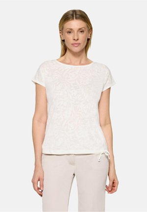 Lyshåret kvinde iført hvid top med korte ærmer og blomsterprint samt lyse beige bukser, stående foran en ensfarvet lys baggrund.