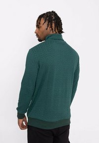 Pull en tricot vert avec un col montant, présentant un motif en losange texturé et des poignets et un ourlet côtelés. Fond uni.