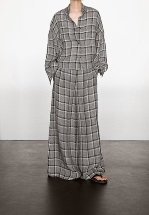 Loose gray and black plaid button-up shirt paired with matching wide-leg pants and black sandals, standing against a plain white wall.