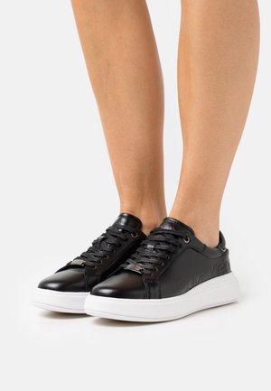 Baskets en cuir noir avec semelles en caoutchouc blanc, dotées de lacets ronds, d'un logo gaufré sur le côté et d'une texture lisse. Design bas classique.