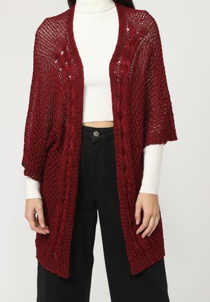 Cárdigan de punto burdeos con patrón de trenzado, abertura frontal y mangas cortas, superpuesto a una blusa blanca de canalé, combinado con pantalones negros.