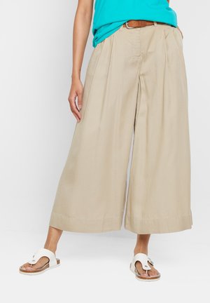 Donna che indossa pantaloni beige a gamba larga e corti, top turchese, cintura marrone e sandali bianchi aperti sulle dita, in piedi su sfondo bianco.
