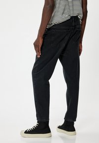 Jeans de mezclilla negra con un corte relajado, que cuentan con bolsillos traseros y puños doblados. Se llevan con zapatos de charol negros y una camisa a rayas.