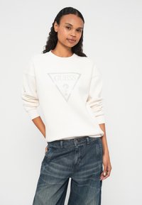 Weißer Pullover mit einem dreieckigen Grafikmotiv und silbernen Buchstaben in der Mitte. Dazu lockere, dunkle Jeans mit sichtbaren Nähten.