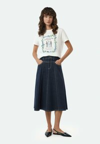Witte t-shirt met kleurrafdruk, gecombineerd met een donkerdenim midi rok met brede plooien en zakken. Zwarte platte schoenen maken de look compleet.