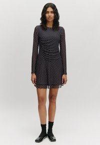 Zwarte jurk met polkadots, lange mouwen, gerimpelde lijfje en doorschijnende rok. Heeft een zachte textuur en een uitlopende zoom. Gedragen met zwarte sokken en schoenen.