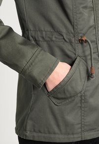 Olivgrön jacka i texturerat tyg, justerbar dragsko i midjan och en sidoficka. Har en figursydd ärmdesign.