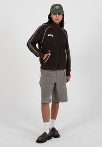 Brun teksturert jakke med glidelås og hvite detaljer, kombinert med lysegrå shorts. Modell har på seg grønn lue og brune sko.