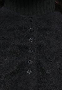 Μαύρο πλεκτό πουλόβερ με ψηλό ραμμένο γιακά, απαλό και χνουδωτό υφασμά, και έξι μαύρα κουμπιά που διατρέχουν το μπροστινό μέρος.