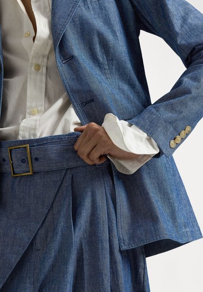 Džínový oblek so štruktúrovaným sako a plisovanými nohavicami, oba v svetlom modrom odtieni. Zvýraznený zlatou prackou na opasku a bielou košeľou pod ním.