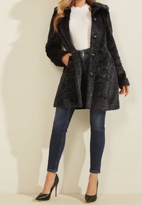 Abrigo de piel sintética negra con un patrón texturizado, botones delanteros y capucha, combinado con jeans de mezclilla oscura y tacones negros.