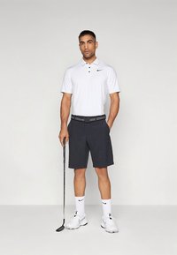 Hvit polo med svart logo, svarte shorts og hvite sportssko. En golfkølle holdt i én hånd. Enkel, avslappet sportsklærstil.