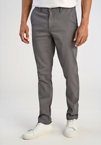 Mies, joka käyttää harmaita slim fit -housuja ja valkoisia lenkkikenkiä, seisoo vaaleataustalla kädet rentona sivuilla.