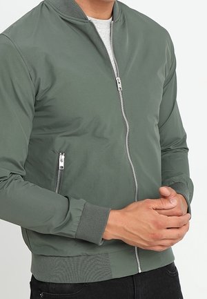 Personne portant une veste bomber verte avec une fermeture éclair à l'avant et une poche zippée, mains croisées devant, sur un fond uni.