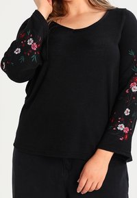 Haut noir à manches longues avec broderie florale rouge et blanche sur les manches. Col en V, coupe décontractée et texture douce.