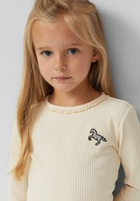 Kreem värvi ribakujuline pikakä sleeve top, millel on sakiline krae ja väike tikand hobuse kujutisega rinnal. Pehme, tekstuuriga kangas.