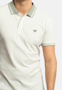 Polo de color claro con un patrón texturizado, un cuello gris en contraste y un pequeño logo verde en el pecho. Mangas cortas.