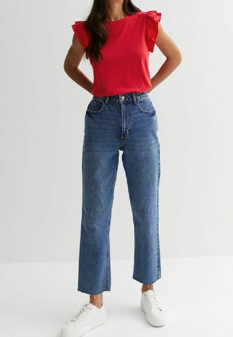 Haut rouge à manches courtes avec des accents de volants, associé à un jean bleu taille haute à jambes droites et fentes latérales, porté avec des baskets blanches.