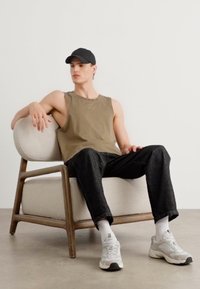 Olivengrønn ermeløs topp, mørk denim-bukse, hvite sokker, grå sneakers. Sittende i en beige polstret stol med trebelte.