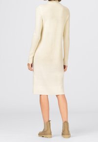 Robe-pull crème longueur genou avec manches longues, poignets et ourlet côtelés, présentant une texture lisse et un design simple. Les bottes sont beige.