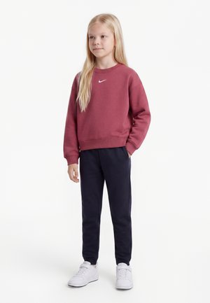 Młoda dziewczyna o długich blond włosach, ubrana w bordową bluzę Nike, ciemne spodnie i białe trampki, stoi z jedną ręką w kieszeni.