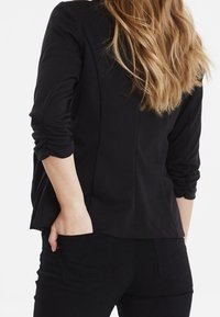 Svart jerseyblazer med en skräddarsydd design, uppvikta ärmar och subtila sömdetaljer, kombinerad med svarta slim-fit byxor.