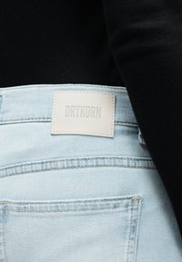Svetlo modre jeans hlače iz jeansa z belo pravokotno blagovno znamko "DRYKORN" nad zadnjim desnim žepom, nošene s črnim topom.