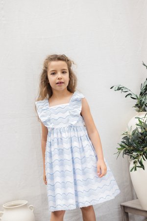 Φόρεμα από ανοιχτό μπλε βαμβάκι με βολάν στους ώμους και κυματιστό σχέδιο· μήκος μέχρι το γόνατο με σούρα στη μέση· συνδυασμένο με ουδέτερο φόντο και φύλλωμα.