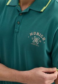 Tyrkysové polo tričko so žltými akcentmi, vybavené tromi gombíkmi, štruktúrovanou tkaninou a vyšitým brandingom "Mníchov 1939" na hrudi.