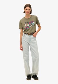 Olijfgroene katoenen T-shirt met kleurrijk bloemdesign en "Superdry" tekst, gecombineerd met lichtgewassen spijkerbroek en zwarte schoenen.