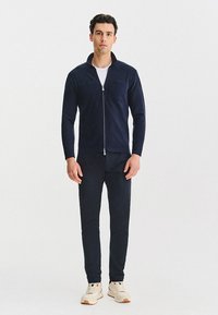 Granatowa kurtka zapinana na zamek z dwoma kieszeniami na piersi, w połączeniu z pasującymi spodniami slim-fit i kremowymi sneakersami. Gładka tekstura, minimalistyczny design.