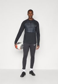 Cămașă neagră cu mânecă lungă și panou căptușit la piept, asortată cu pantaloni negri tapered și pantofi sport negri. Ține o minge de fotbal.