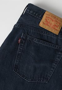 Σκούρο μπλε τζιν Levi's με έμφαση στην πίσω τσέπη και καφέ δερμάτινη ετικέτα της μάρκας στη μέση, σε ανοιχτό γκρι φόντο.