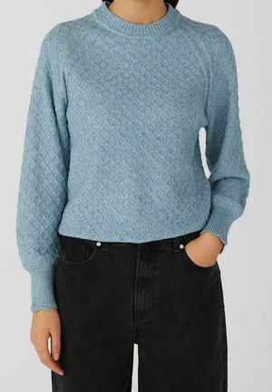 Persoon die een lichtblauwe, gestructureerde gebreide trui met lange mouwen en zwarte, hooggetailleerde jeans draagt, met één hand in de zak.