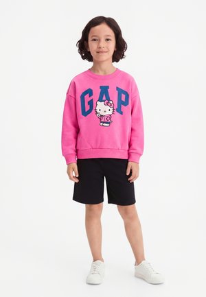 Kind met donker haar draagt een roze sweatshirt met Hello Kitty en blauwe letters GAP, zwarte korte broek en witte sneakers, staand tegen een witte achtergrond.