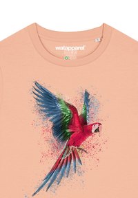Perziken t-shirt met een kleurrijke papegaai graphic met blauwe en groene vleugels, gedetailleerde texturen en een spetterverf effect.