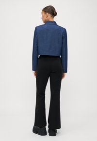 Kraftig blå denimjakke med strukturerte skuldre og kontraststing, parret med svarte flagrende bukser og klumpete svarte sko.