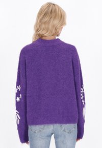 Violetti neulepaita, jossa on pörröinen rakenne ja valkoista kukkakirjontaa hihoissa. Lyhyt leikkaus ja pyöreä pääntie.