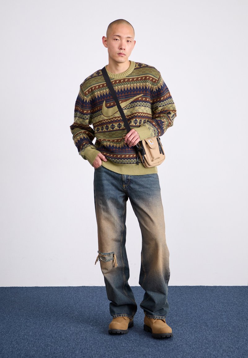 Pull en tricot vert avec des motifs multicolores, jeans en denim déchirés et chaussures beiges. Petit sac à épaule beige avec des bretelles noires.