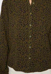 Camisa verde oliva de botões, com um padrão de manchas pretas, textura suave e um design ligeiramente curto, com botões visíveis.