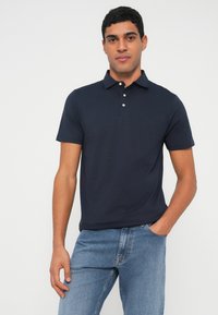 Vīrietis tērpies tumši zilā īsroku polo krekliņā ar trim pogām un gaiši zilos džinsos, stāv ar vienu roku kabatā.