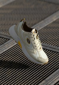 Zapatilla deportiva color crema con suela texturizada, detalles en amarillo y un cuello acolchado. Presenta un parche con logo y un diseño de cordones.