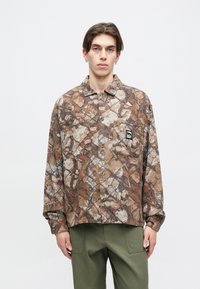 Barna és bézs kamuflázs ing, gombos kialakítással, hosszú ujjakkal és mellzsebbel, amely egy fekete logópatcshoz tartozik.