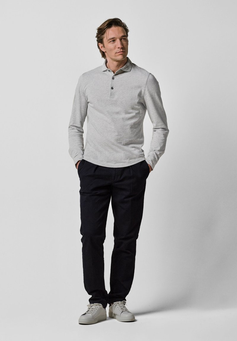 Graues Langarm-Polohemd mit drei Knöpfen, kombiniert mit dunklen Hosen und weißen Sneakers. Minimalistisches Design und glatte Textur.