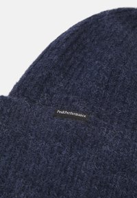 Marine gebreide beanie van zacht garen. Heeft een geribbelde textuur en een klein zwart label met witte tekst dat "PeakPerformance" leest.