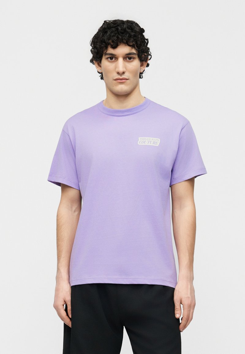 Νεαρός άνδρας με κοντά σγουρά μαλλιά που φοράει μια ανοιχτό μωβ μπλούζα Versace Jeans Couture και μαύρο παντελόνι, στέκεται μπροστά σε απλό φόντο.